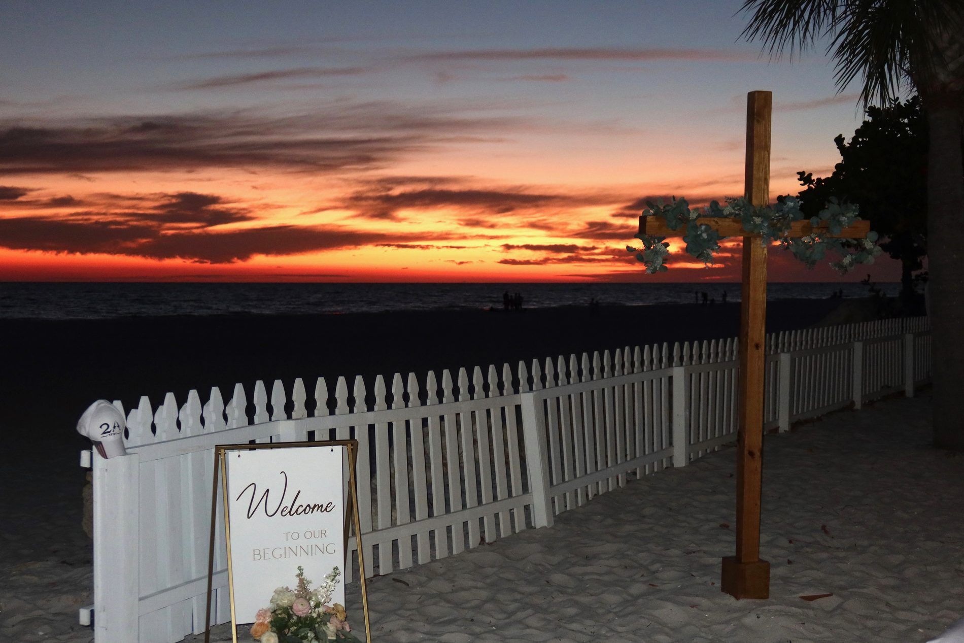 The stained wooden cross dressed with eucalyptus on a Gulf beach at sunset, beside a welcome sign and white picket fence